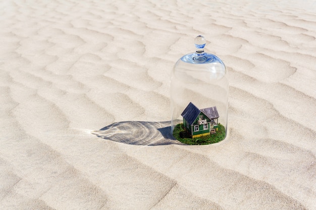 Casa de cartón de juguete con césped verde protegido por una campana de cúpula de vidrio entre un desierto de arena sin vida