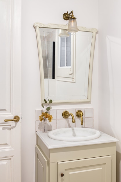 Foto casa de banho decorada na cor bege com pequena torneira de bronze para pia e espelho vintage.