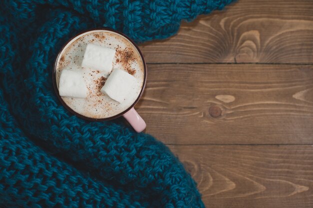 Casa de inverno aconchegante. Uma xícara de chocolate com marshmallows e um cachecol de malha azul em uma mesa de madeira marrom. Lugar para texto.