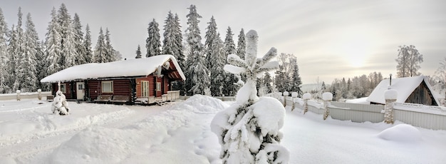 Casa de toras de madeira manchada de cor marrom escuro, com telhado coberto de neve na borda da floresta de abetos de inverno.