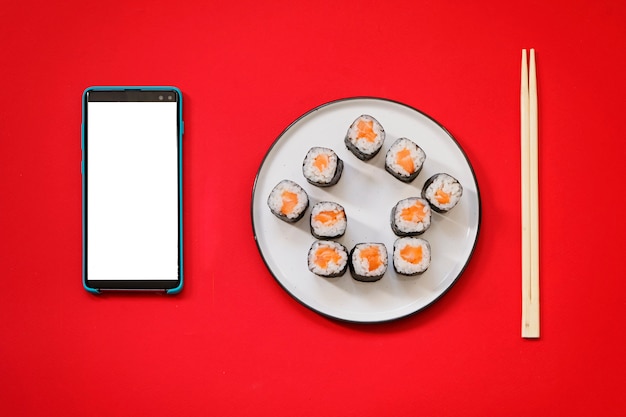 Casa forma sushi rolos com pauzinhos e smartphone em fundo vermelho