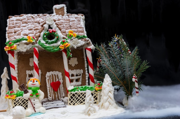 Casa grande de pão de mel caseiro coberto de neve, um galho de árvore de Natal e um boneco de neve de mástique de açúcar em fundo escuro. Maquete para ofertas sazonais e cartão postal de férias