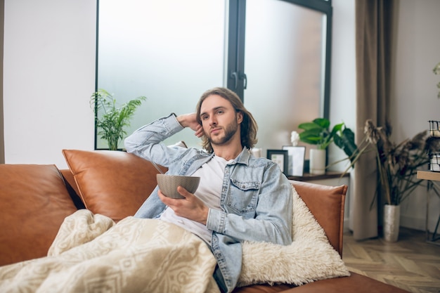 En casa. Hombre de pelo largo en camisa de jeans descansando en casa