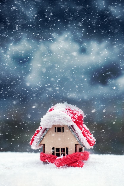 Una casa de juguete de madera en la nieve contra el fondo de un cielo oscuro y nublado en invierno durante las nevadas está cubierta con un sombrero y envuelta con una bufanda El concepto de preservación del calor