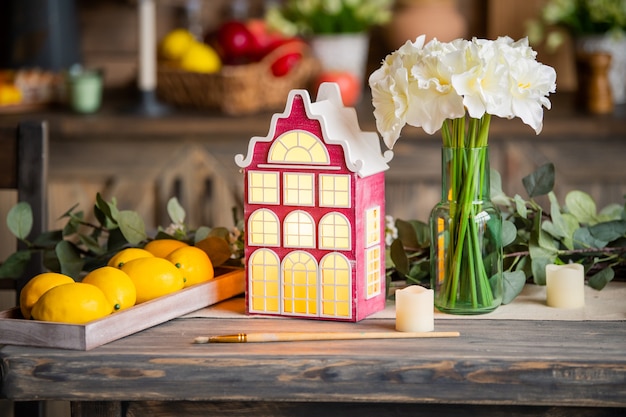 La casa de la lámpara decorativa de juguete se encuentra en una mesa de madera entre flores de primavera y elementos interiores.