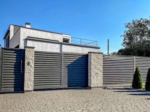 Una casa particular gris de dos pisos escondida detrás de una cerca con puertas marrones corredizas automáticas y un área cercana a la casa contra un cielo azul.