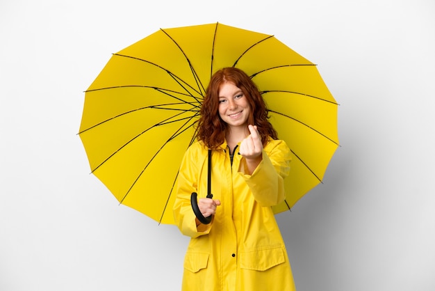Casaco à prova de chuva e guarda-chuva da menina ruiva adolescente isolado no fundo branco fazendo gesto de dinheiro