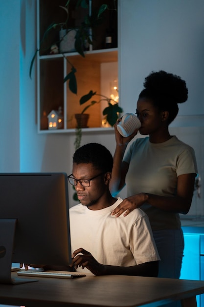 Casado con un adicto al trabajo. Joven africana con una taza de té parada detrás del hombre mientras codifica en la computadora y trabaja de forma remota desde casa hasta altas horas de la noche, la esposa brinda apoyo al marido trabajador