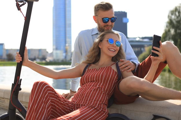 Casal adorável se divertindo dirigindo uma scooter elétrica, fazendo uma pausa na direção, sentado na margem do rio, tirando uma selfie,