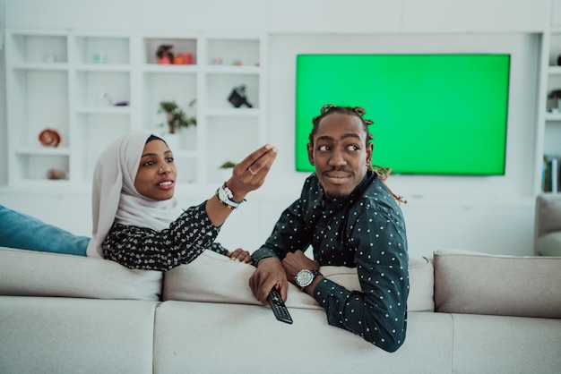 Casal africano sentado no sofá assistindo tv juntos mulher de tela verde cromática usando hijab islâmico