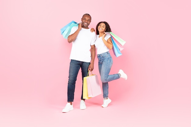 Casal afro-americano feliz com sacolas de compras posando em fundo de estúdio rosa espaço livre de tiro completo