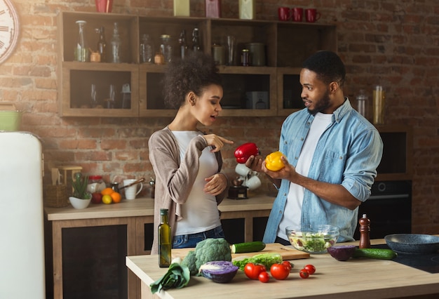 Casal afro-americano preparando salada de legumes saudável com pimenta no interior do loft