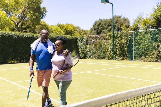 Casal afro-americano sênior feliz com raquetes de tênis caminhando em uma quadra de grama ensolarada, espaço de cópia. Estilo de vida sênior, aposentadoria, esporte, verão, fitness, hobbies e atividades de lazer.