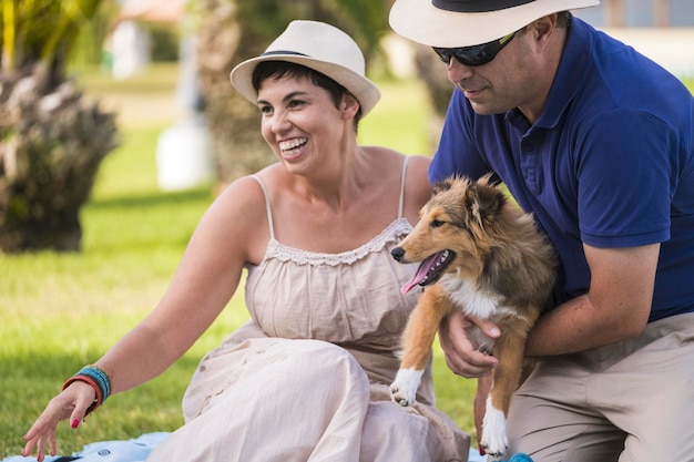 Casal alegre caucasiano de meia-idade em atividade de lazer ao ar livre, brincando com um belo cachorrinho de shetland sentado em um campo de grama verde e se divertindo todos juntos