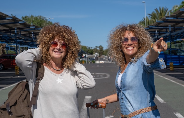 Casal alegre de mulheres encaracoladas com bagagem e mochila pronta para viajar, sorrindo, olhando para a câmera