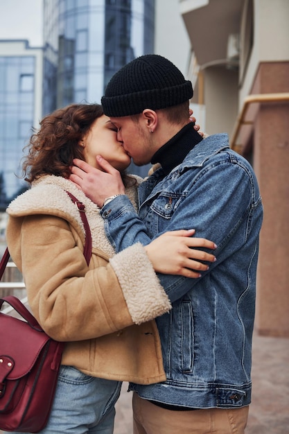 Casal alegre em roupas quentes casuais beijando ao ar livre na cidade perto de prédio comercial