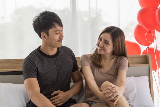 Casal, amor e conceito de dia dos namorados. retrato de dois homem asiático sorridente e mulher olhando um para o outro rosto e sentado na cama no quarto com balão vermelho.