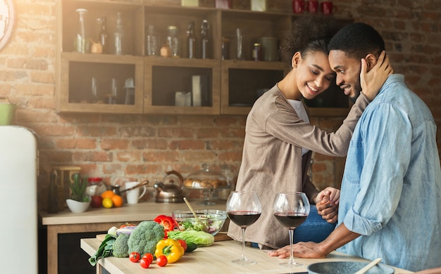 Casal amoroso afro-americano, abraçando e bebendo vinho na cozinha. Família preparando o jantar