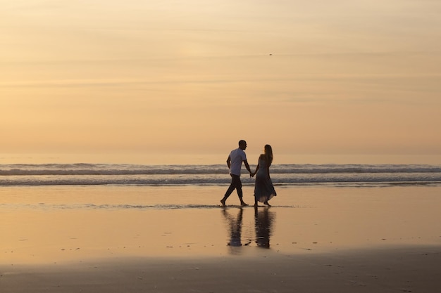 Casal apaixonado andando na praia ao pôr do sol. homem e mulher em roupas casuais passeando pela água ao entardecer. amor, família, conceito de natureza