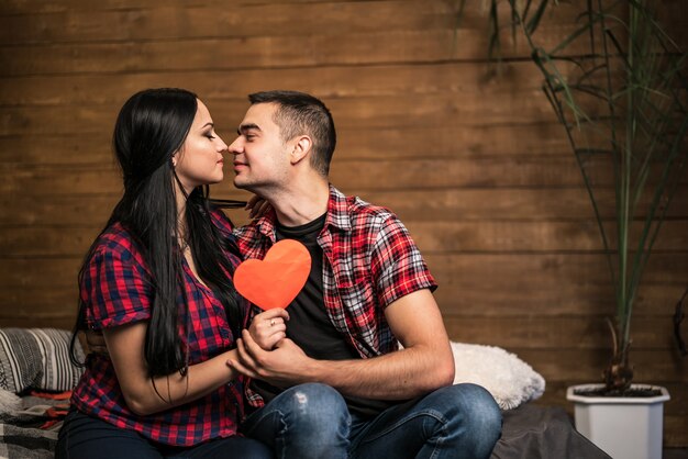 Foto casal apaixonado beijando com cartão de coração de papel vermelho em casa. beijo sensual de um lindo casal deitado na cama enquanto comemorava o dia dos namorados.