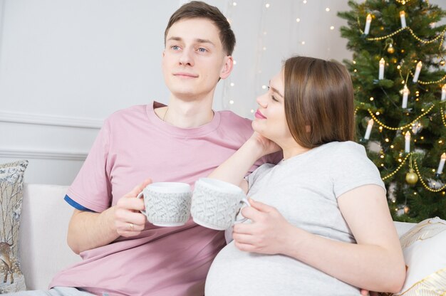 Casal apaixonado celebra o Natal. Mulher grávida segurando o copo com bebida quente. Árvore de Natal em segundo plano. Boas festas.