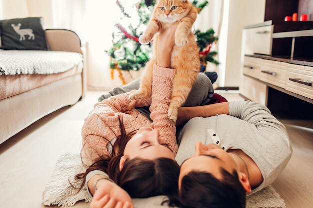 Casal apaixonado, deitado perto da árvore de Natal e brincando com o gato em casa. Homem e mulher levantando animal de estimação nas mãos. Família feliz