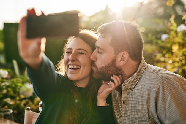 Foto casal apaixonado e selfie em um pátio por pessoas felizes se beijando e relaxando ao pôr do sol juntos telefone da família e homem beijando mulher para foto enquanto está sentado em um quintal sorrindo e amando em casa