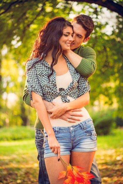 Casal apaixonado em pé nas folhas caídas do outono, o cara abraçando e beijando uma garota bonita no pescoço.