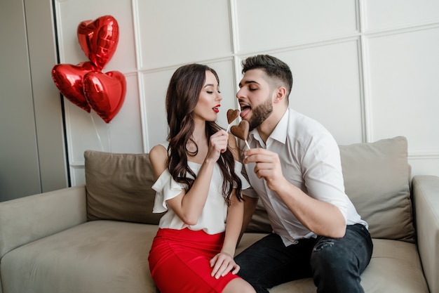 Casal apaixonado homem e mulher com coração doce em forma de bombons de chocolate e balões vermelhos, sentado no sofá em casa