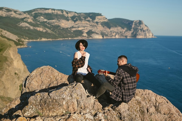 Casal apaixonado no topo de uma montanha com vista para o mar homem toca violão uma mulher de chapéu canta