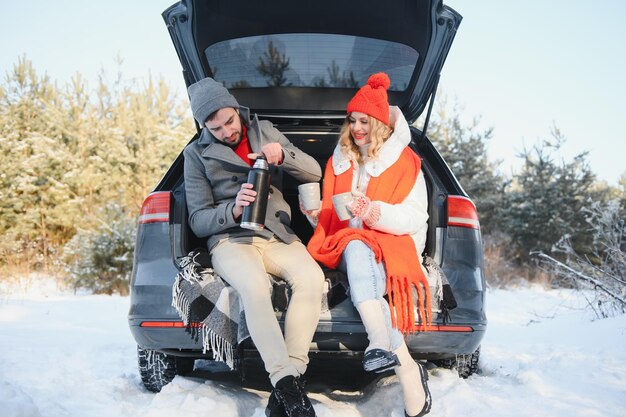 Casal apaixonado, sentado na mala do carro, bebendo chá quente na floresta de inverno nevado e conversando. Pessoas relaxando ao ar livre durante a viagem. Dia dos namorados