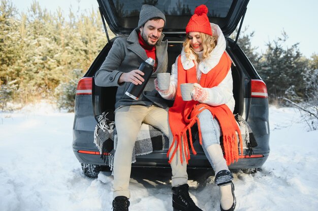 Casal apaixonado, sentado na mala do carro, bebendo chá quente na floresta de inverno nevado e conversando. Pessoas relaxando ao ar livre durante a viagem. Dia dos namorados