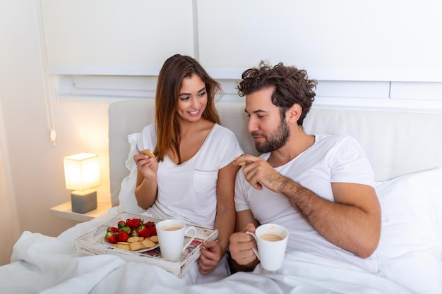 Casal apaixonado tomando café da manhã na cama Jovem casal caucasiano tomando café da manhã romântico na cama Feminino e masculino duas xícaras de frutas de café e biscoitos coloridos