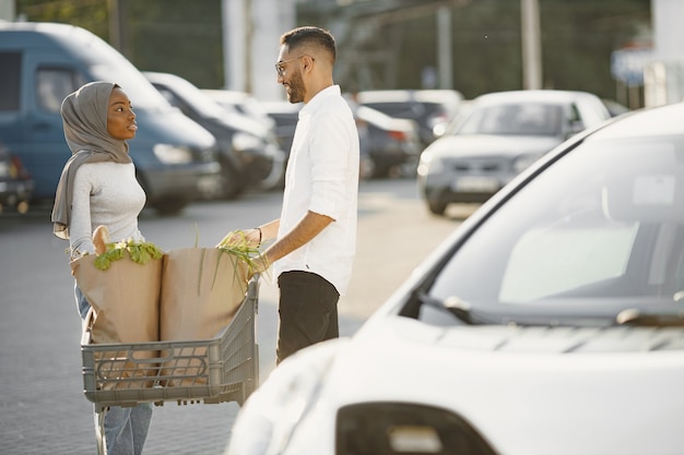 Casal árabe africano fica com mantimentos perto de carro elétrico. carregando carro elétrico no posto de gasolina elétrico