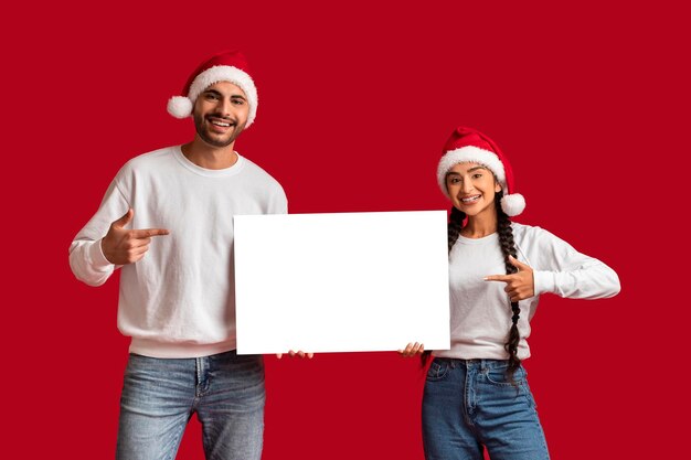 Casal árabe feliz em chapéus de Papai Noel apontando para cartaz em branco nas mãos