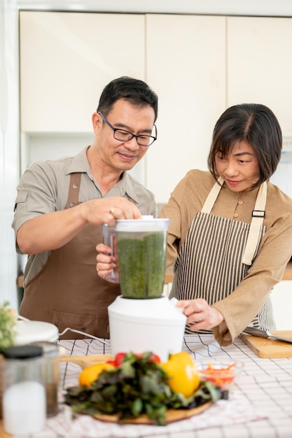 Casal asiático adulto feliz está fazendo smoothie verde saudável na cozinha juntos usando um liquidificador