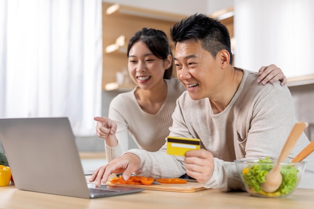 Casal asiático animado fazendo pedido on-line usando laptop na cozinha