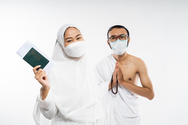 Casal asiático em roupas de ihram e máscara segurando um passaporte com gesto de salam em fundo isolado