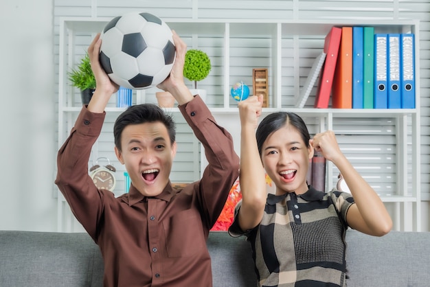 Casal asiático jovem bonito olhando surpreso enquanto está sentado no sofá e assistindo jogo de futebol na tv em casa.