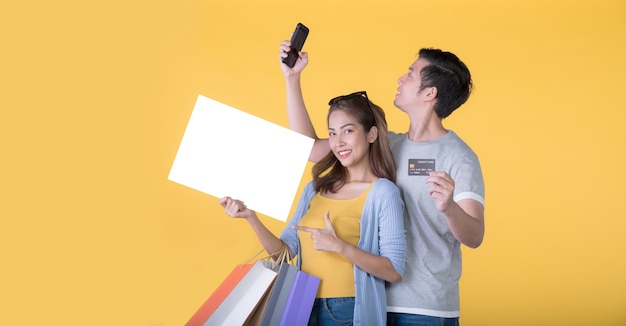 Casal asiático segurando outdoor em branco com cartão de crédito e sacolas de compras e smartphone isolados em fundo amarelo