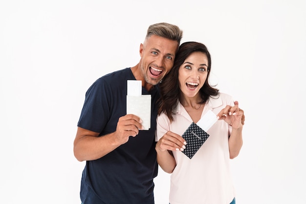 Casal atraente alegre, vestindo roupa casual, em pé, isolado na parede branca, mostrando passaportes com passagens aéreas