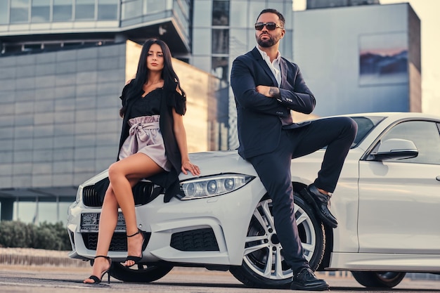 Casal atraente bem vestido, apoiando-se em um carro de luxo ao ar livre contra o arranha-céu.