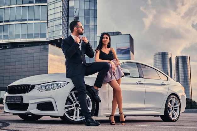 Casal atraente bem vestido, apoiando-se em um carro de luxo ao ar livre contra o arranha-céu.