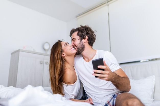 Casal atraente da geração do milênio usando smartphone, deitado na cama juntos, namorado e namorada felizes, verificando as notícias das redes sociais pela manhã, jovem mostrando um novo aplicativo para celular