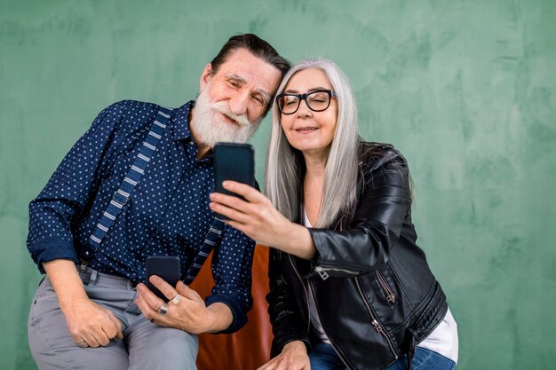 Casal atraente e elegante, homem barbudo e senhora de cabelos grisalhos, sentados juntos na cadeira vermelha e usando os aplicativos de telefone, olhando para a tela do smartphone e sorrindo