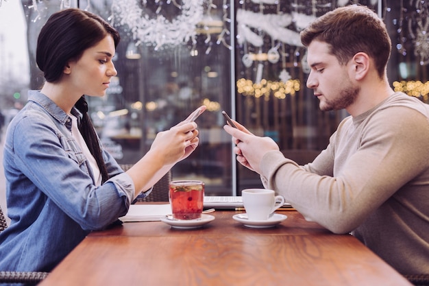 Casal atraente pensativo e sério se ignorando enquanto olha para o telefone e se sentam na frente um do outro