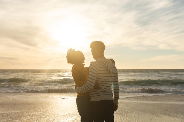 Casal birracial romântico abraçando em pé na praia contra o céu durante o pôr do sol