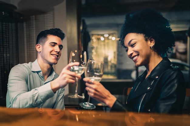 Casal birracial romântico brindando vinho branco sentado em restaurante