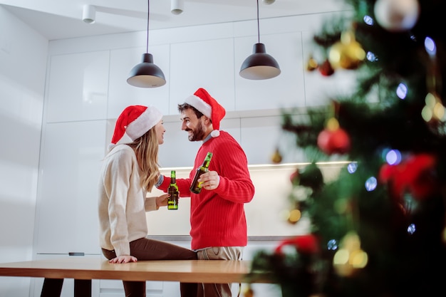 Casal bonito com chapéus de Papai Noel na cabeça, bebendo cerveja e conversando na cozinha