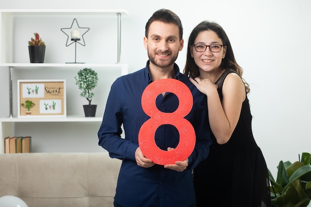 Casal bonito sorridente segurando oito figuras vermelhas em pé na sala de estar em março, dia internacional da mulher